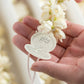 Hand holding a white leaf-shaped object with intricate design against a blurred floral background