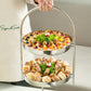 Two-tiered metal tray with assorted food items held by a person against a neutral background