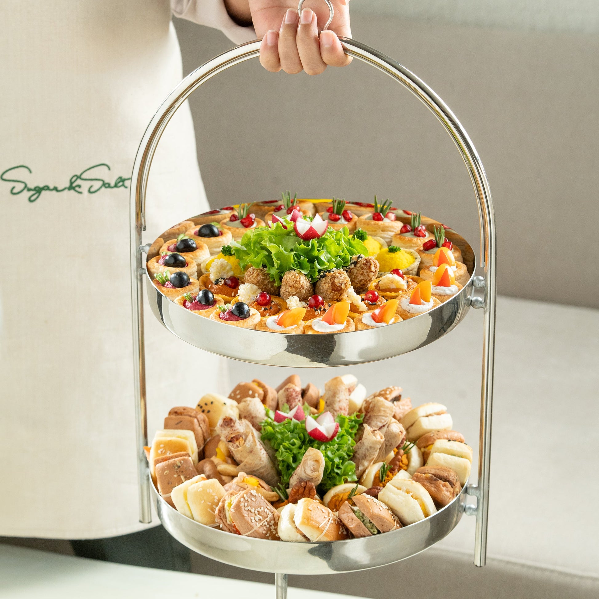Two-tiered metal tray with assorted food items held by a person against a neutral background
